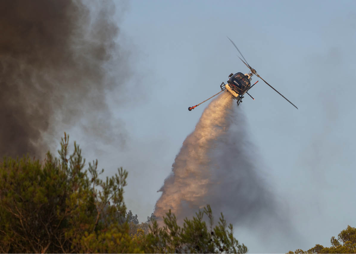 A helicopter is releasing a large plume of water or fire retardant over a forested area with smoke rising nearby. Description generated by AI