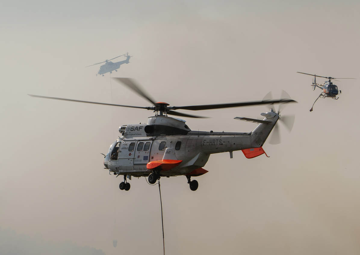 A helicopter with a rope hanging below is flying in the foreground with two other helicopters visible in the background against a hazy sky. Description generated by AI
