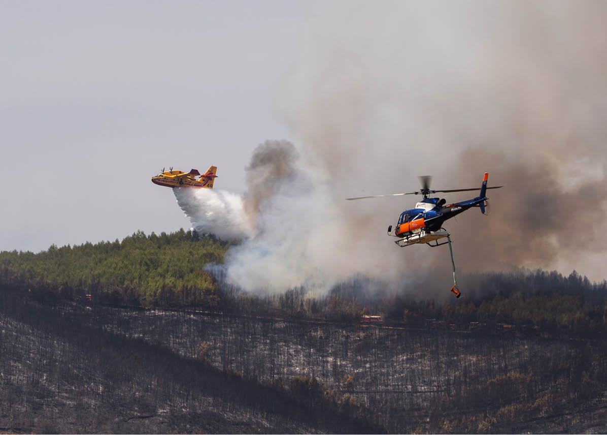 A helicopter and a plane are dropping water over a forest fire with smoke rising from the burned area below. Description generated by AI
