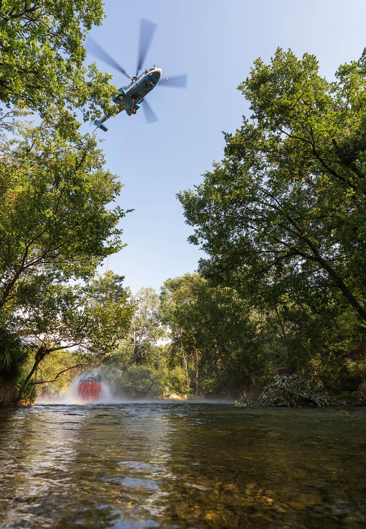 A helicopter is hovering above a river, using a long cable to carry a large bucket that is scooping water from the river surrounded by green trees. Description generated by AI