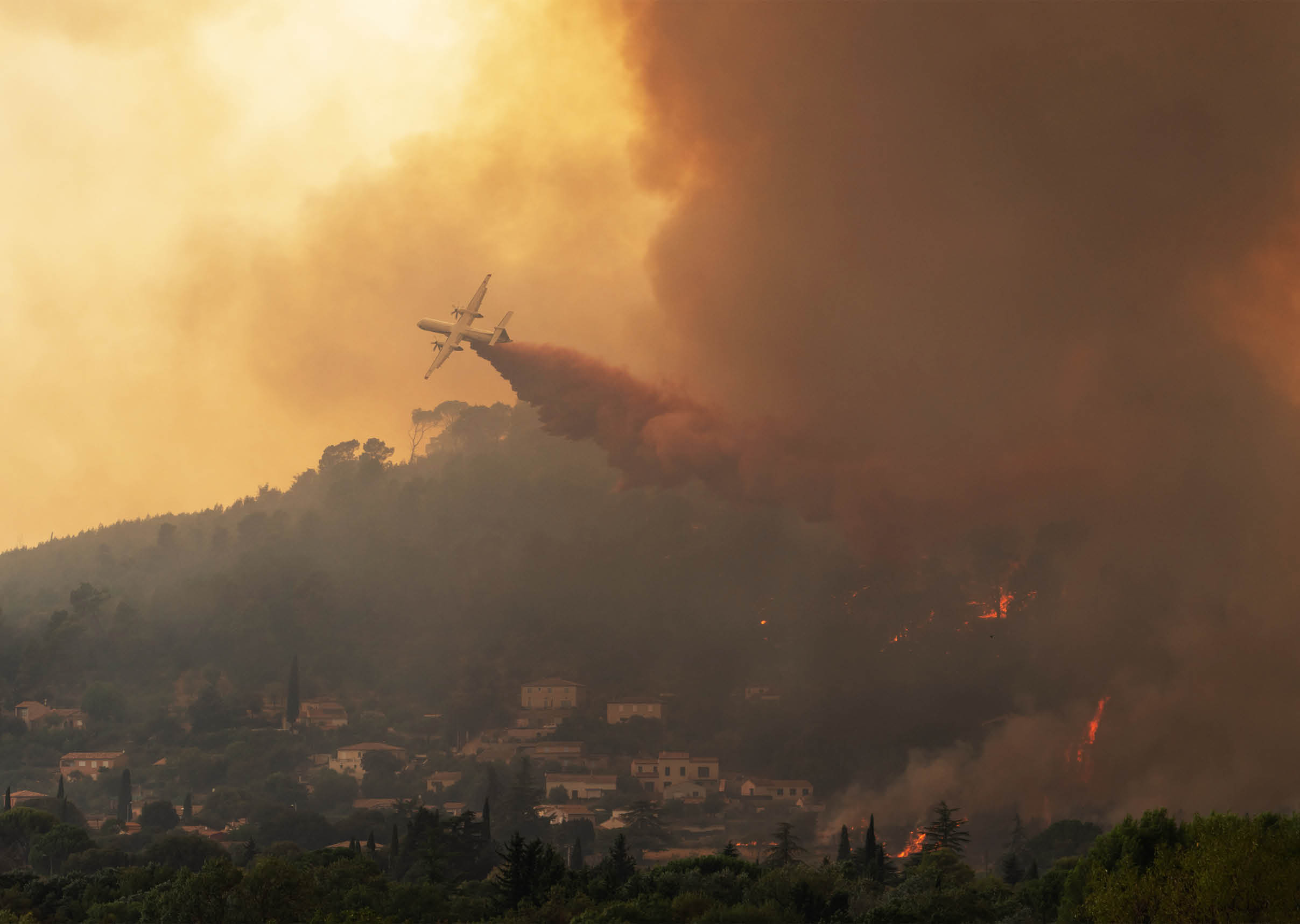 An airplane is releasing fire retardant over a forested hillside engulfed in thick smoke and flames from a wildfire. Description generated by AI