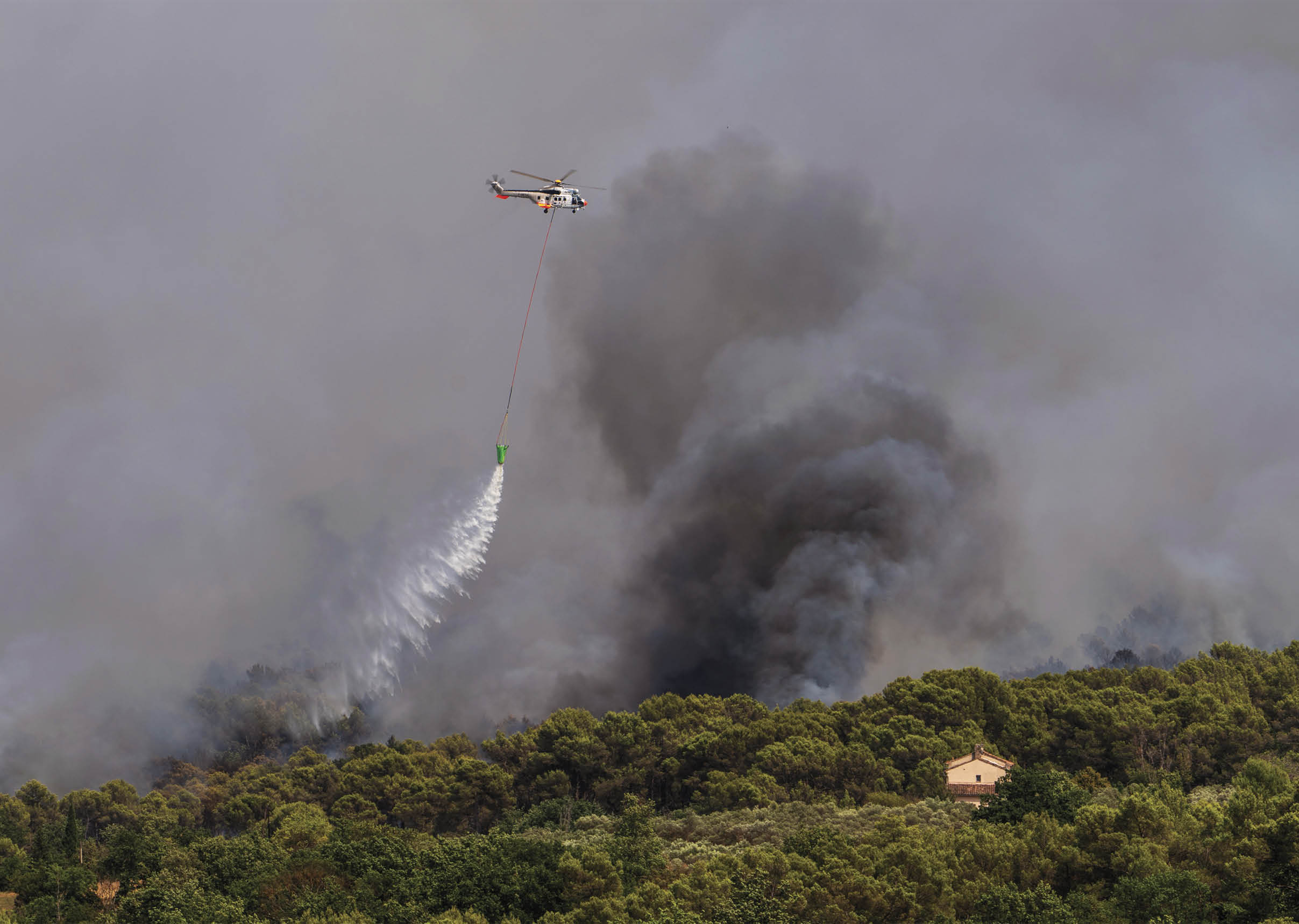 A helicopter is releasing water over a forest fire with thick smoke rising behind the trees near a small house. Description generated by AI