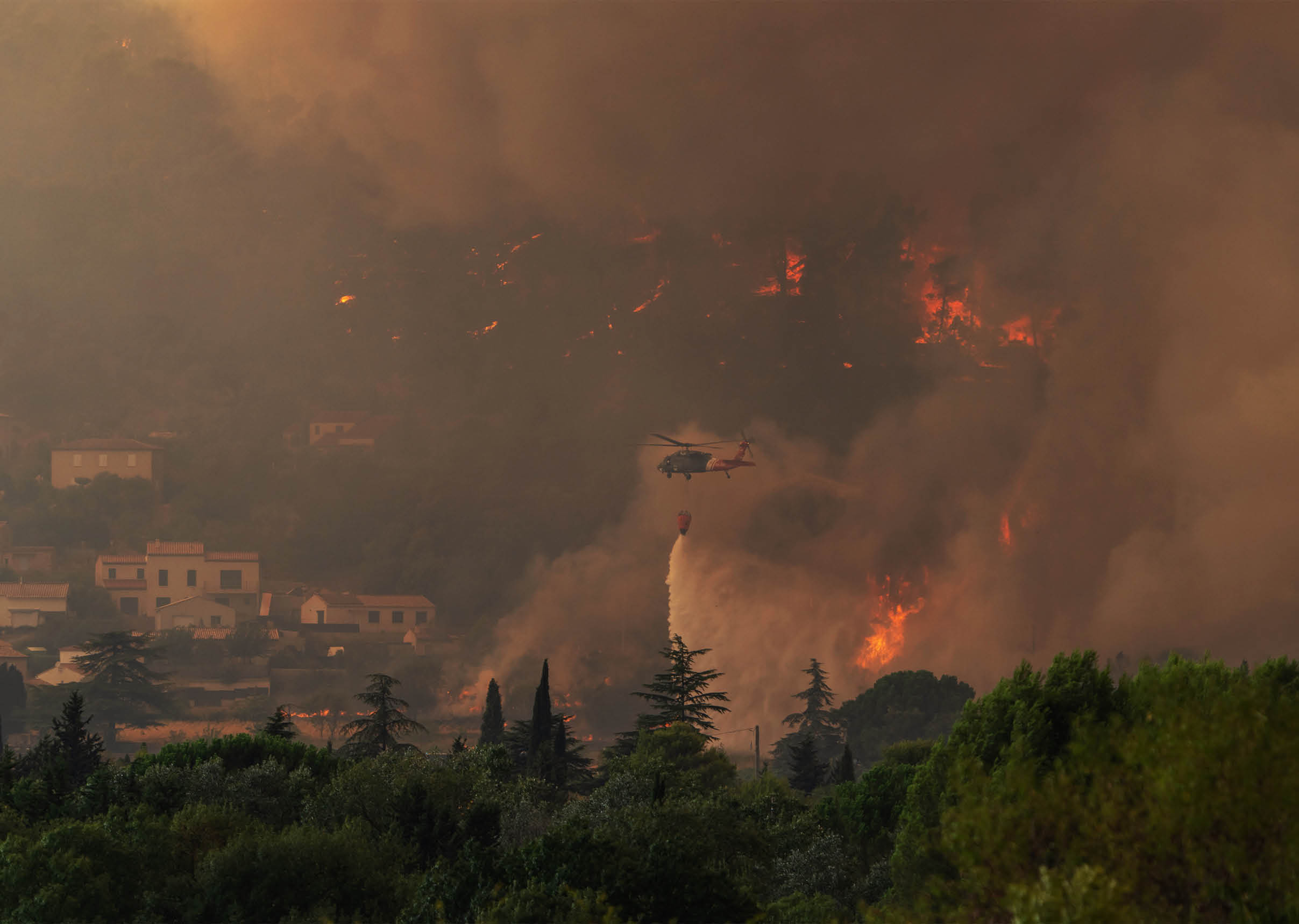 A helicopter is dropping water over a large wildfire burning near a residential area surrounded by trees. Description generated by AI