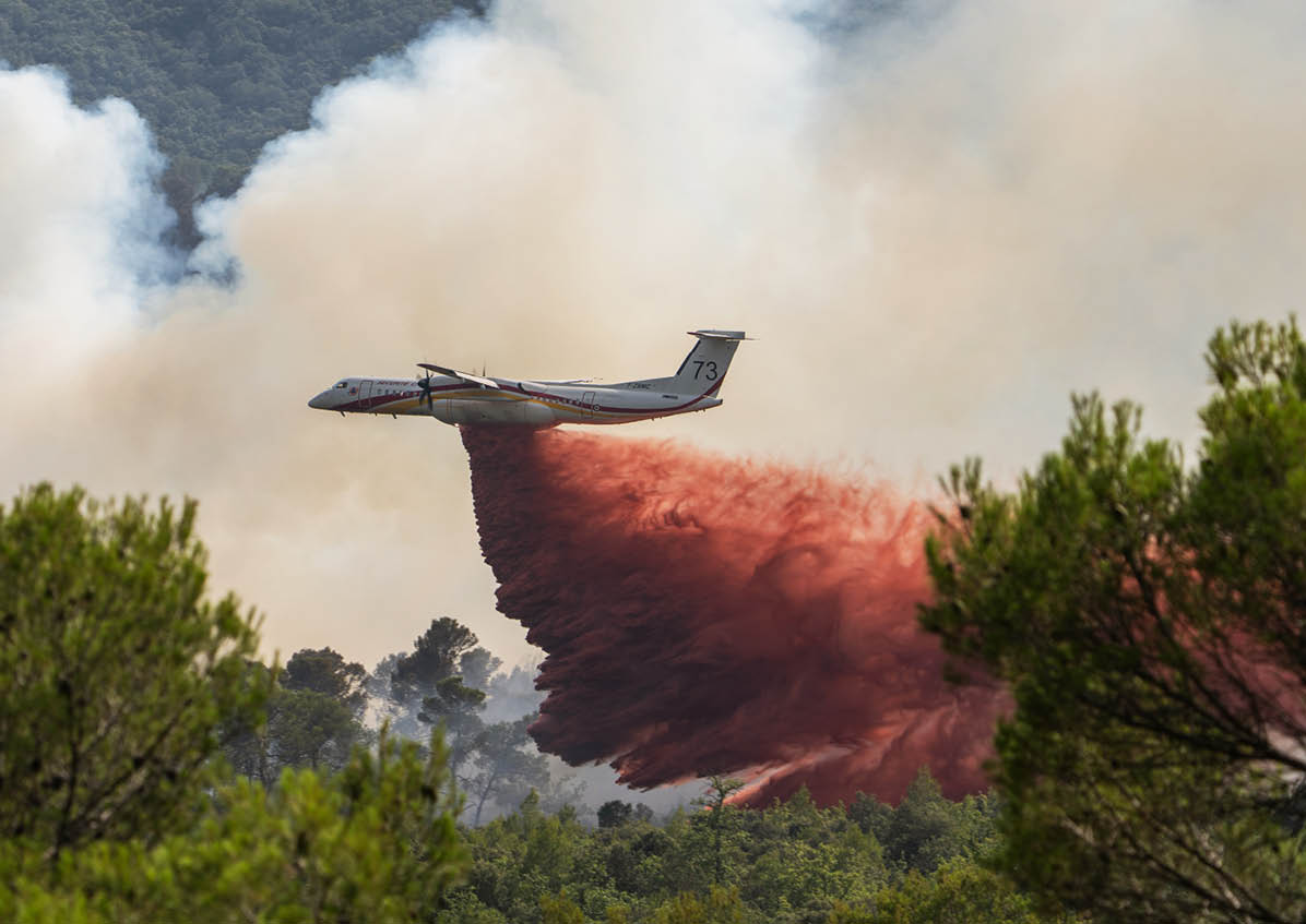 An aircraft is releasing a large plume of red fire retardant over a forested area to combat a wildfire. Description generated by AI
