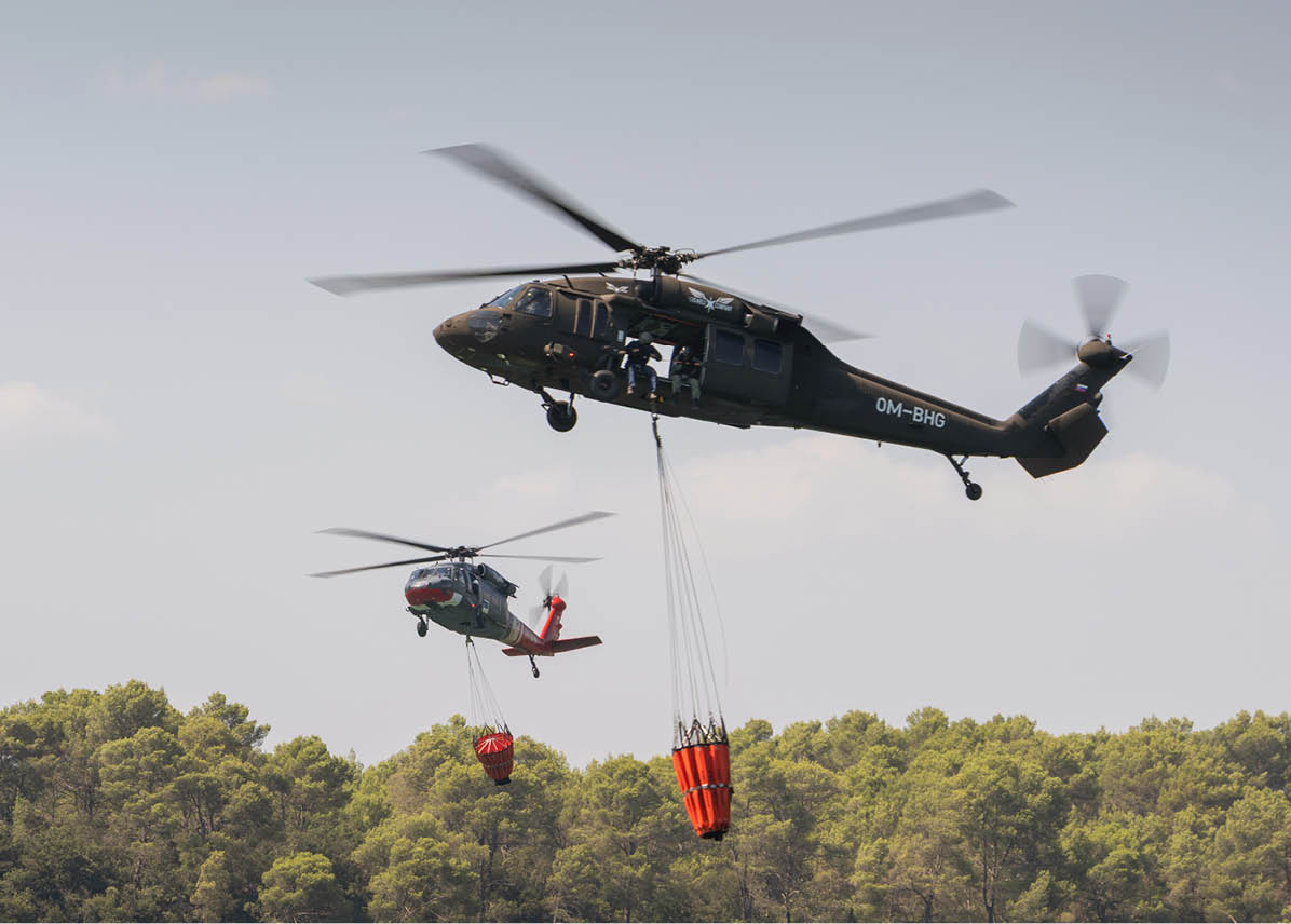 Two helicopters are flying above a forested area, each carrying a large bucket suspended by cables, likely for firefighting purposes. Description generated by AI