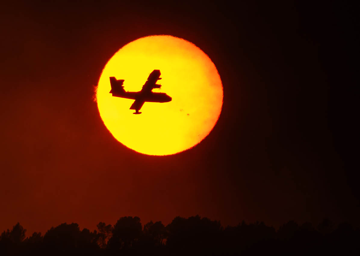 A silhouette of an airplane flying in front of a large, bright orange sun with a dark treeline below. Description generated by AI
