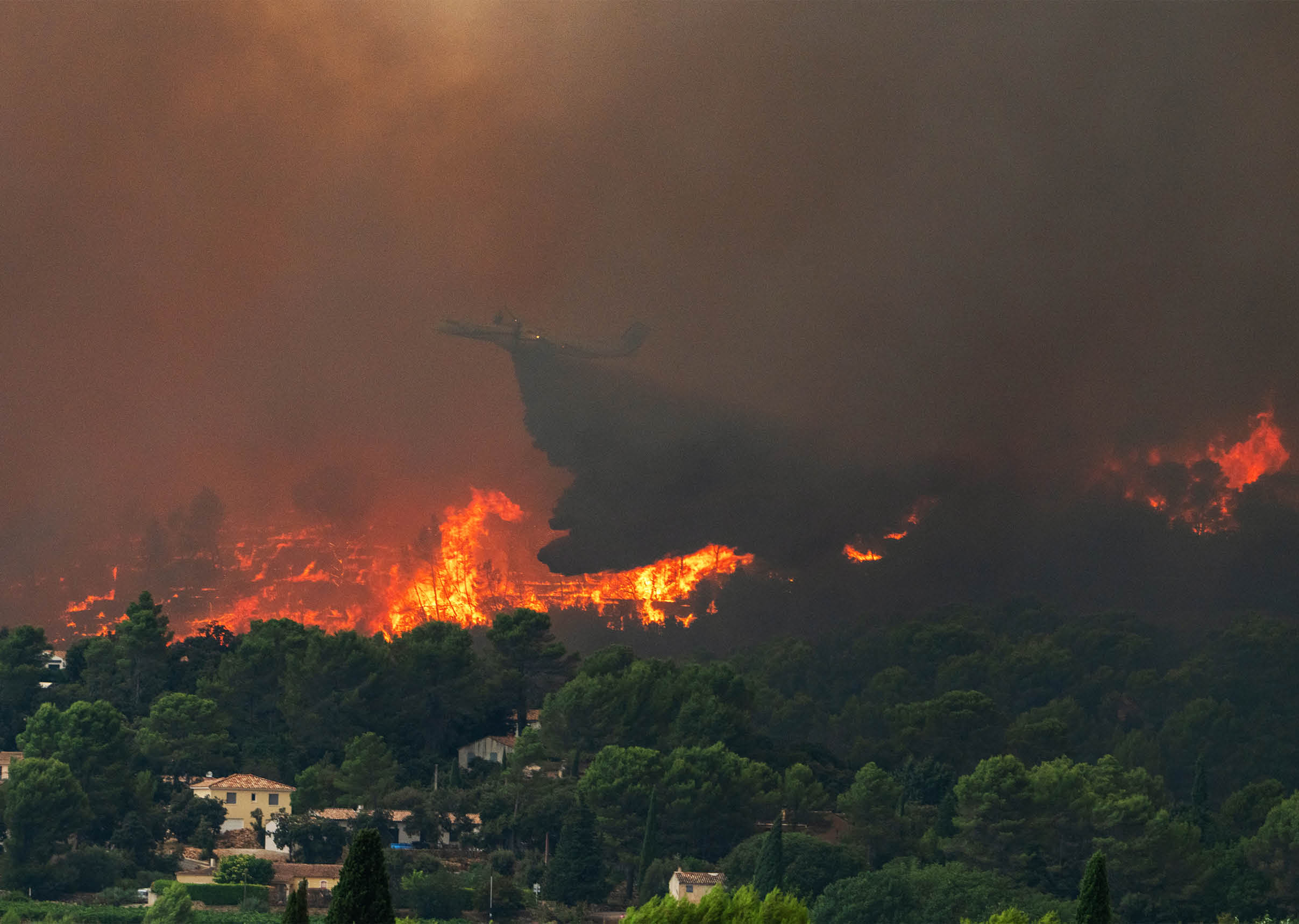 A large wildfire is burning through a forested area near houses with thick smoke rising and a firefighting plane flying above the flames. Description generated by AI