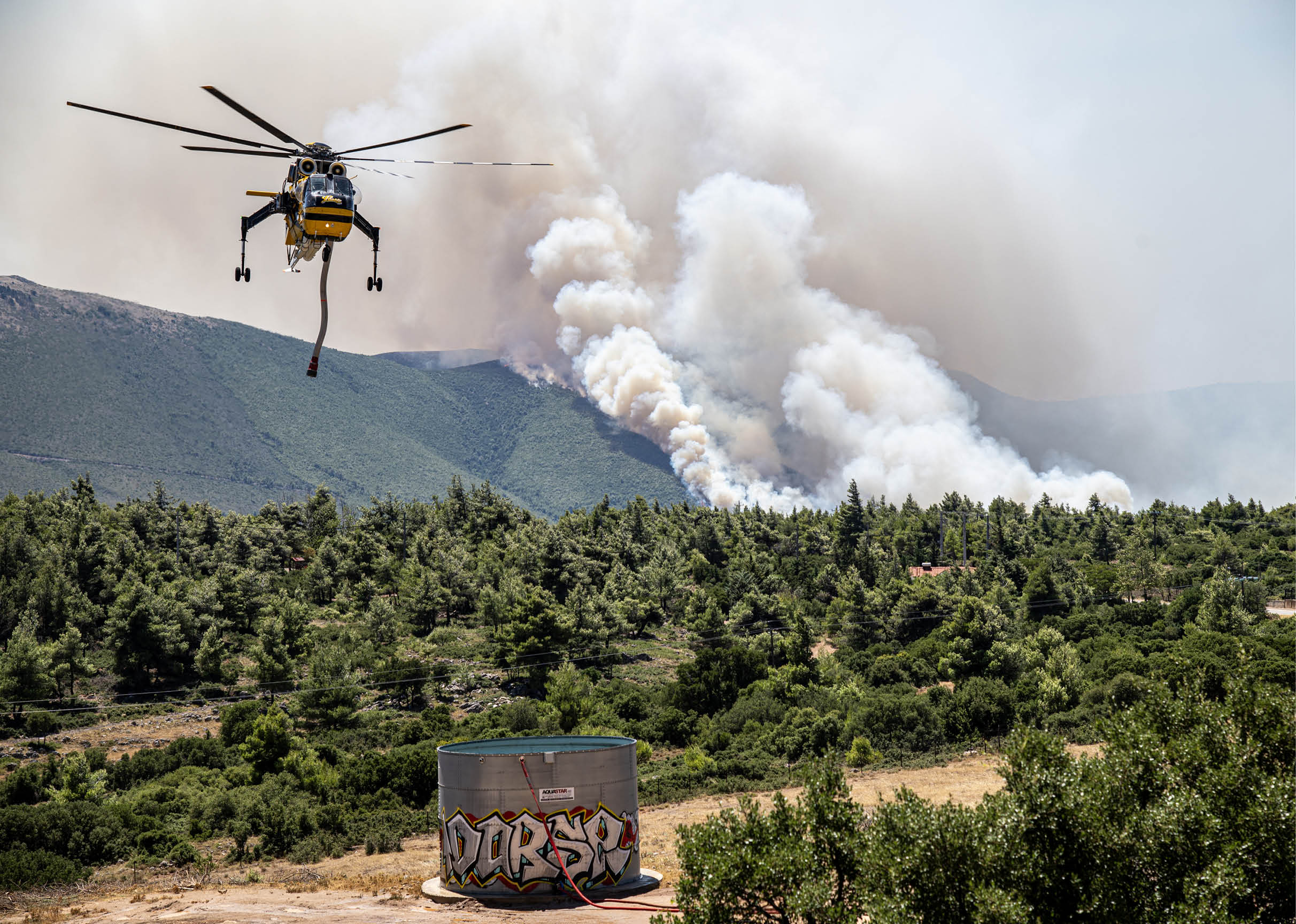 A helicopter is flying over a forested area carrying a bucket while smoke rises from a wildfire on the hillside in the background. Description generated by AI