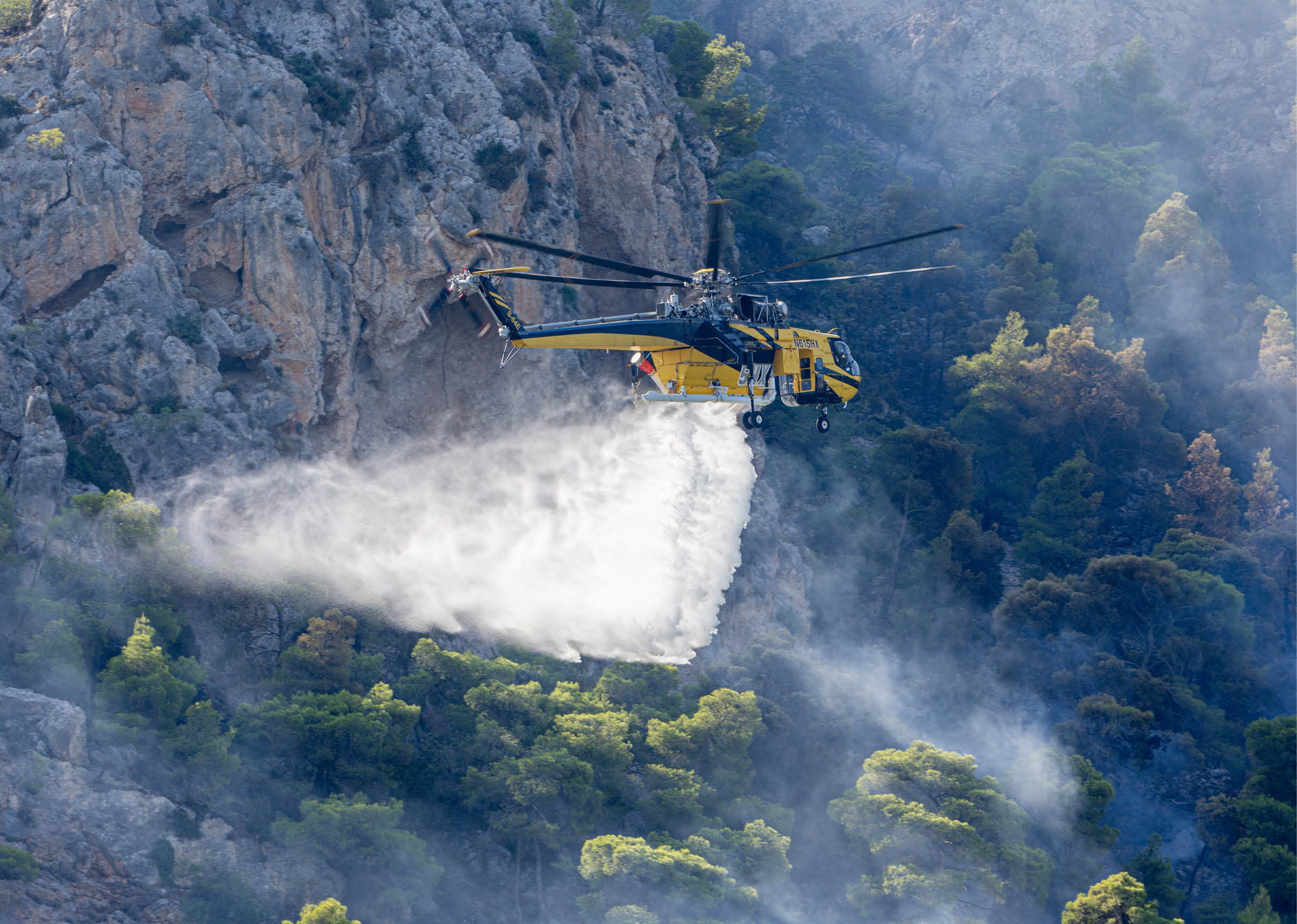 A yellow helicopter is releasing a large spray of water over a forested area near rocky cliffs to extinguish a wildfire. Description generated by AI