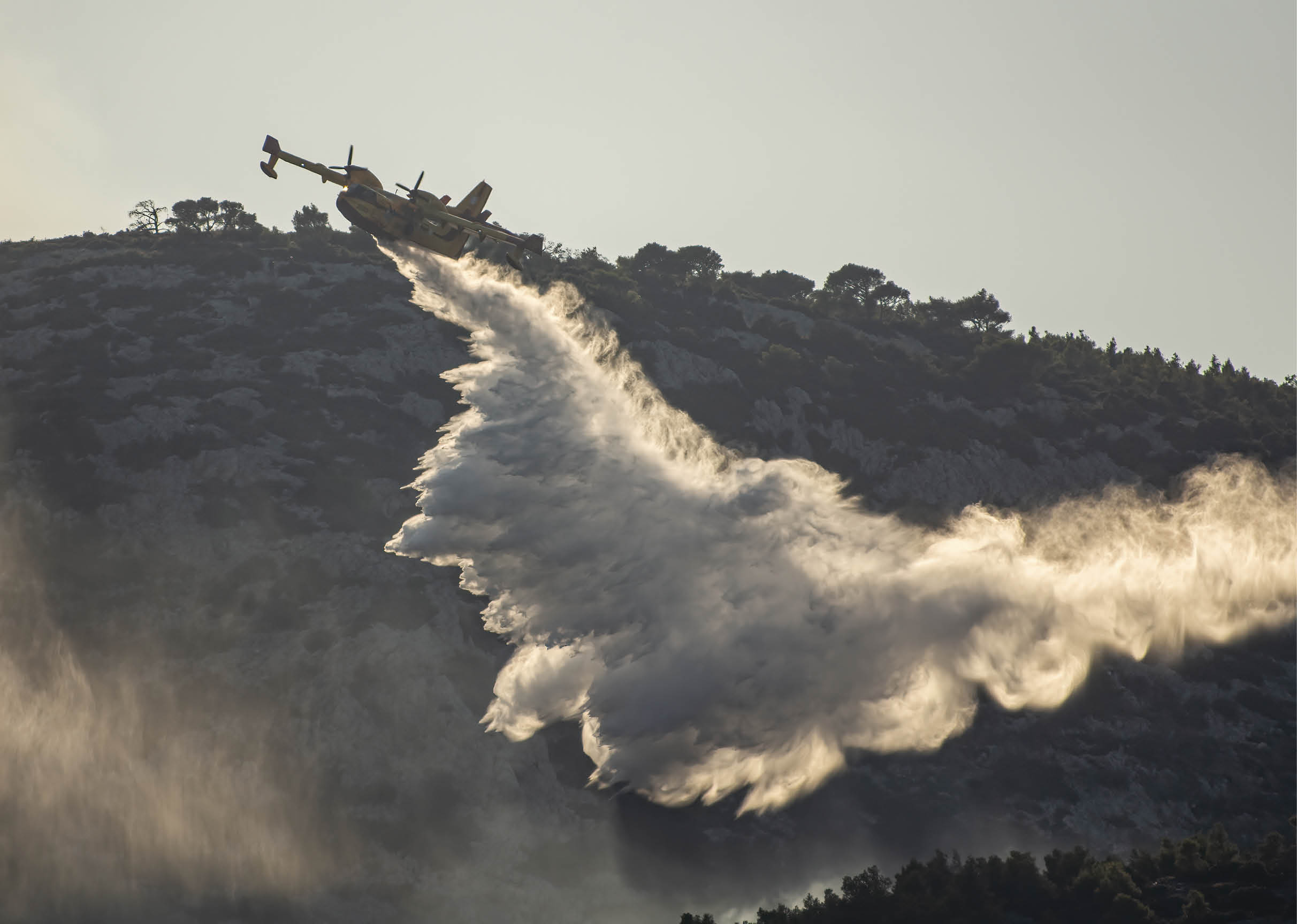 A firefighting airplane is releasing a large plume of water or fire retardant over a forested mountainous area. Description generated by AI