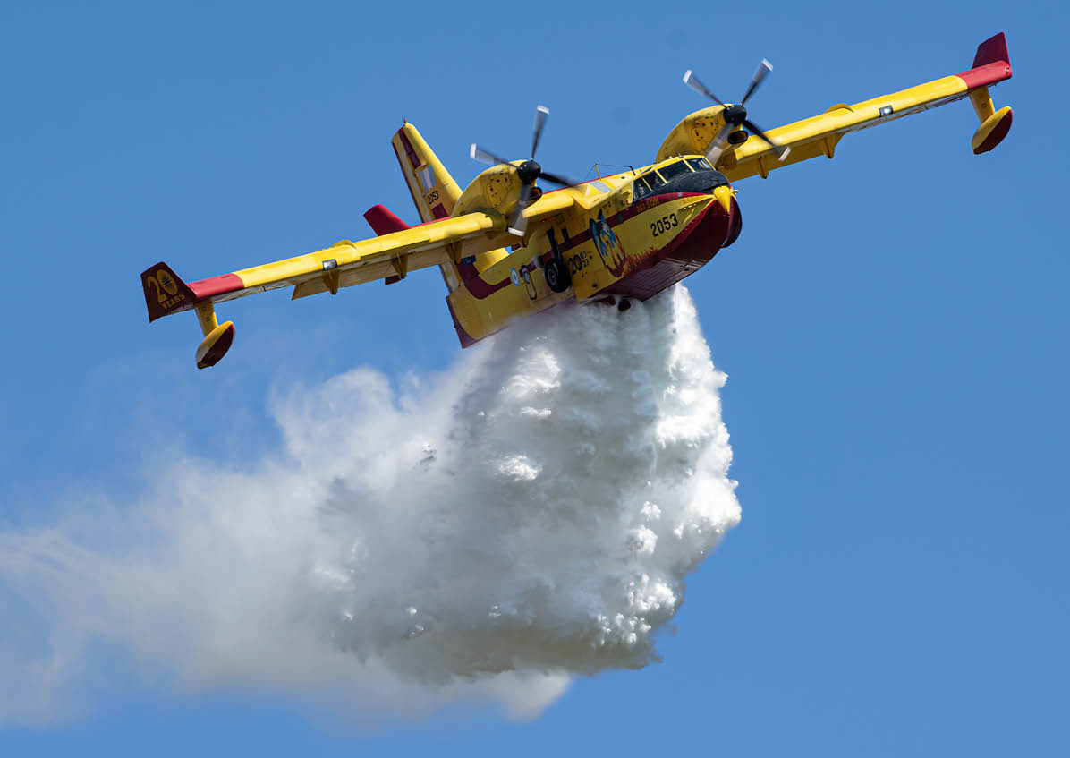 A yellow and red firefighting airplane is releasing a large plume of water or fire retardant against a clear blue sky. Description generated by AI