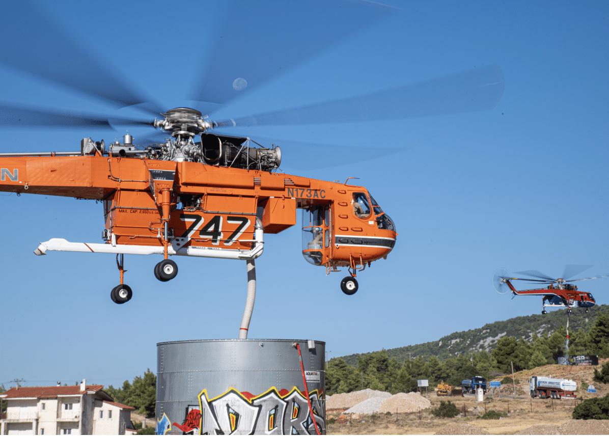 An orange helicopter with the number 747 is hovering above a large cylindrical container with graffiti, with a clear blue sky and a smaller helicopter in the background near a rural area. Description generated by AI