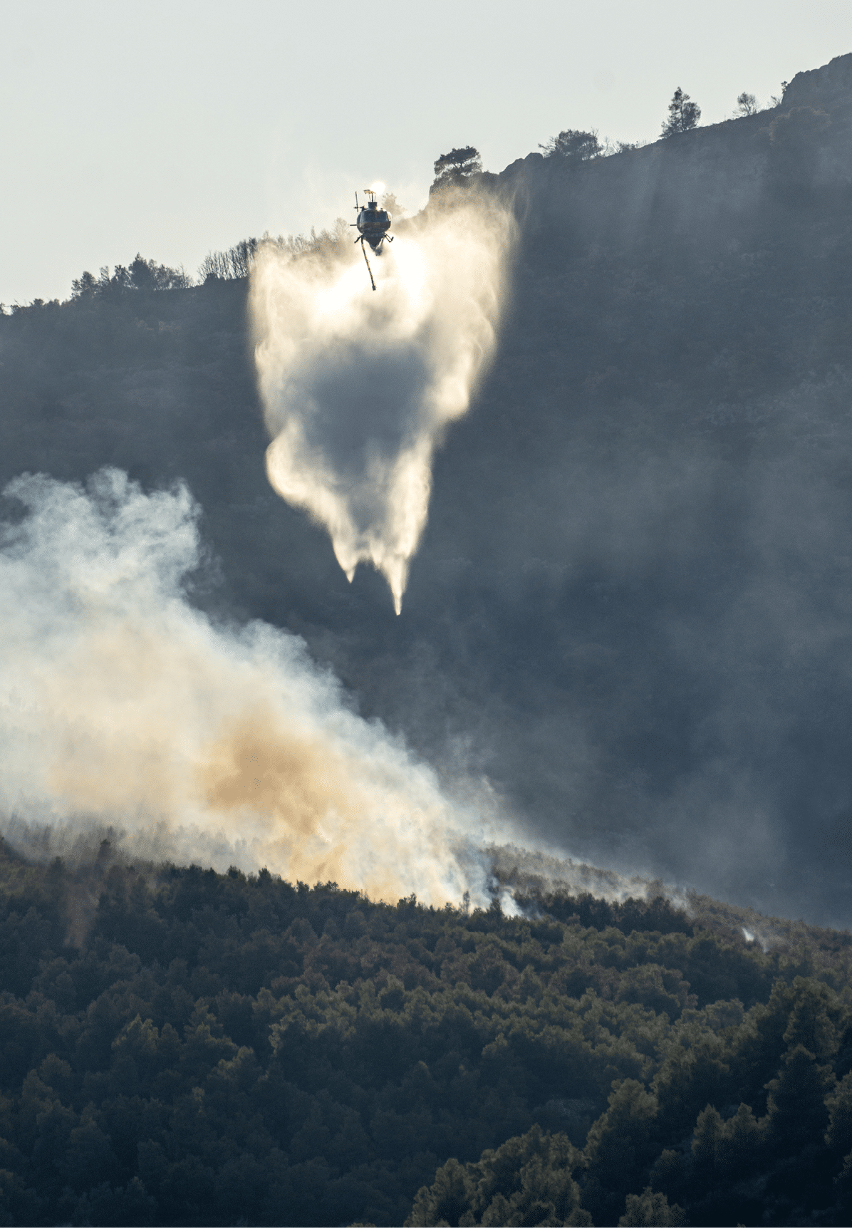 A helicopter is releasing water or fire retardant over a forest fire on a mountainside with smoke rising from the trees. Description generated by AI