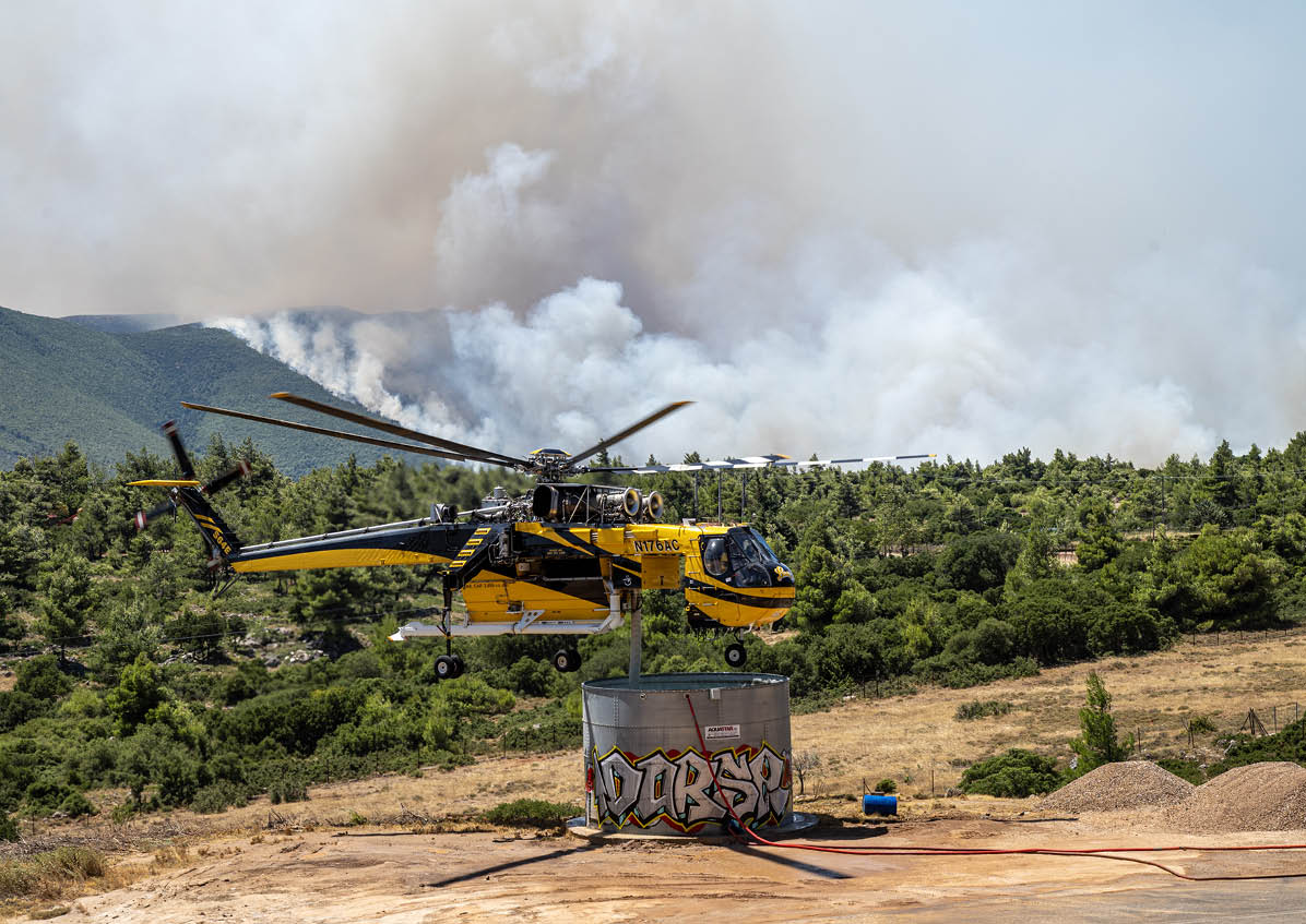 A yellow and black firefighting helicopter is hovering above a large water tank with smoke rising from a forest fire in the background. Description generated by AI