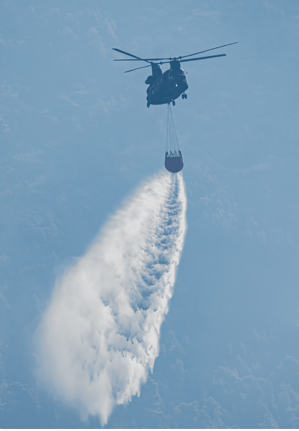 A helicopter is flying in the sky carrying a large bucket that is releasing a stream of water, likely for firefighting purposes. Description generated by AI