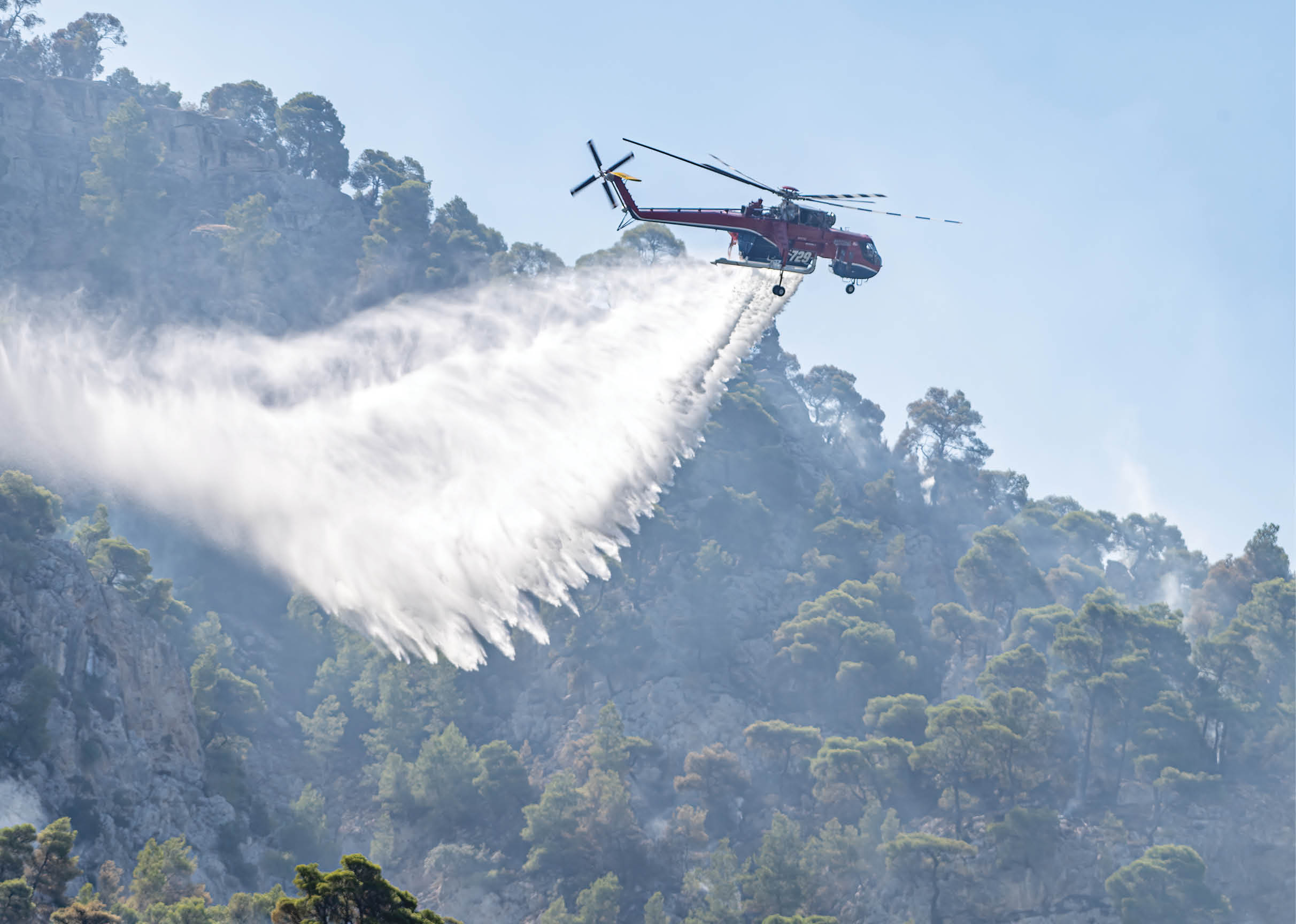 A helicopter is releasing a large spray of water over a forested hillside to extinguish a fire. Description generated by AI