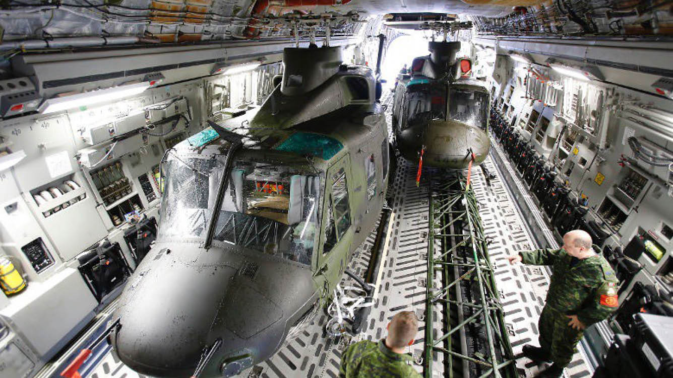 Two helicopters are parked inside a hangar, with one being loaded with supplies. Two men are standing nearby, one of them wearing a camouflage uniform. AI generated content