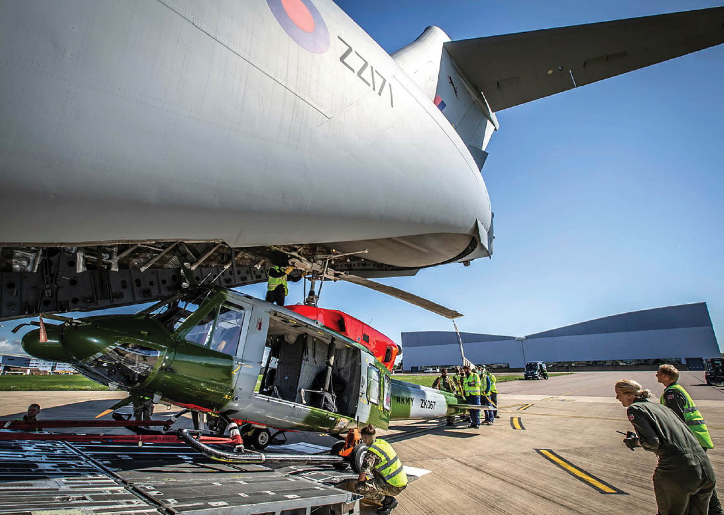 A large airplane has its back opened, revealing a helicopter inside. Several people are working on the scene, with some standing near the helicopter and others near the airplane. The scene appears to be taking place on an airport runway. AI generated content