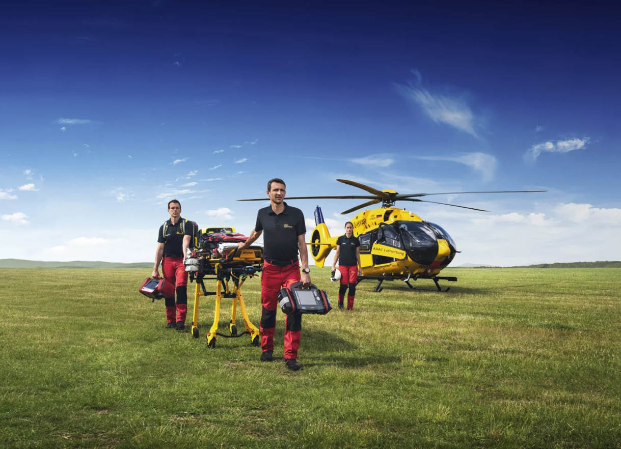 Two men standing in a field with a yellow helicopter in the background. Description generated by AI