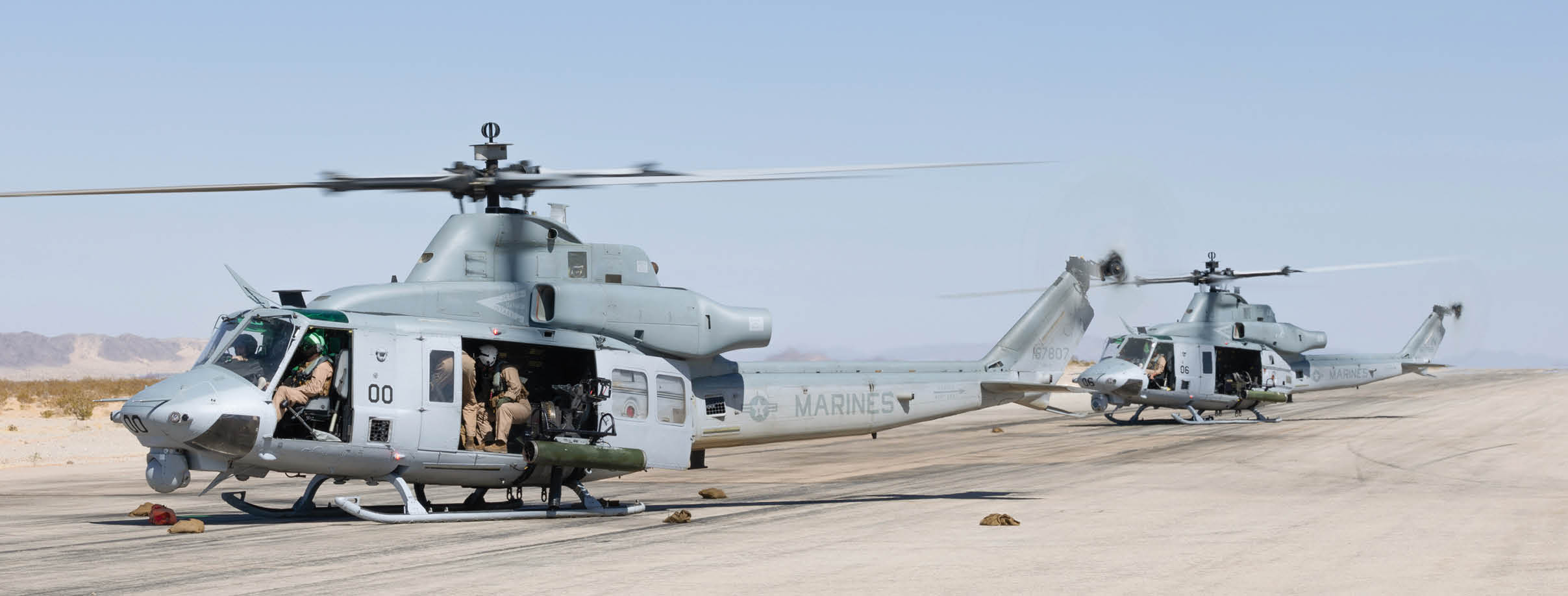 Two military helicopters are parked on a desert runway with several people sitting inside and near the open doors. Description generated by AI