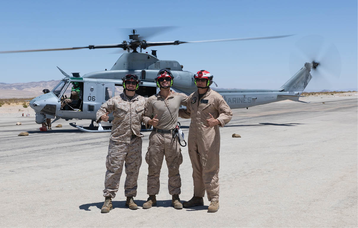 A person wearing a helmet and goggles stands next to a helicopter with another person inside, set against a clear blue sky and desert landscape. Description generated by AI