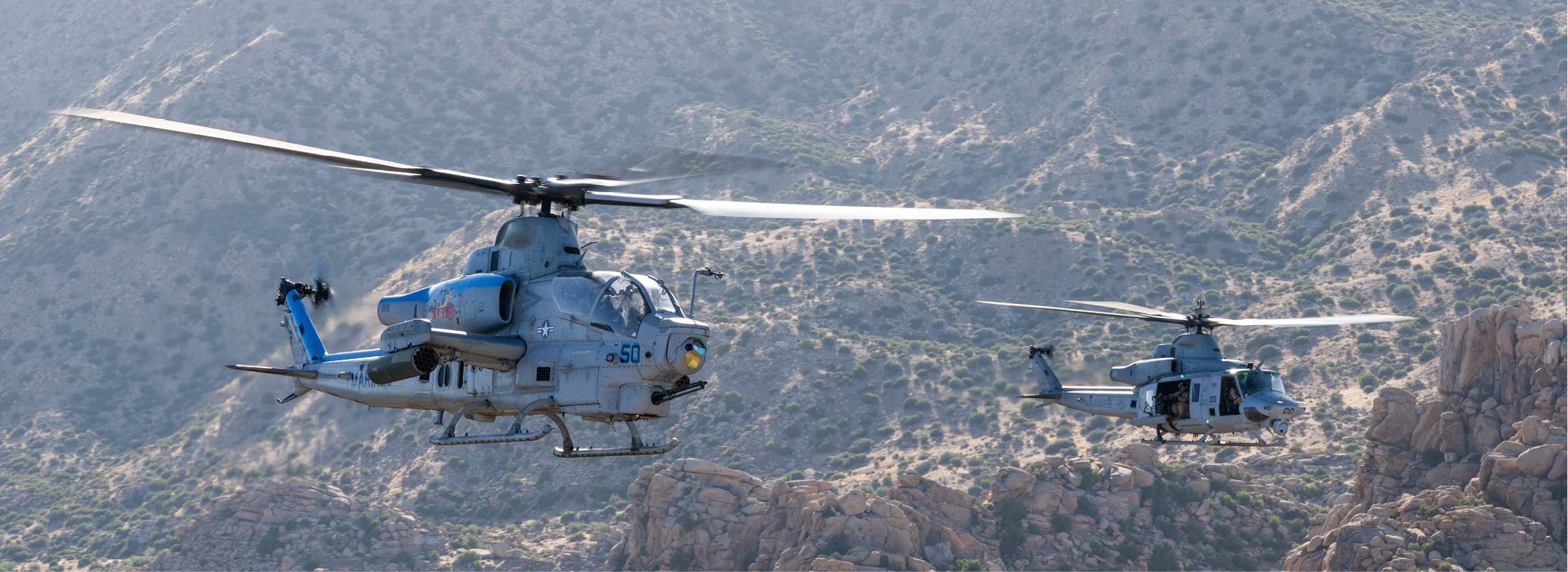 Two military helicopters are flying over a rocky mountainous landscape. Description generated by AI