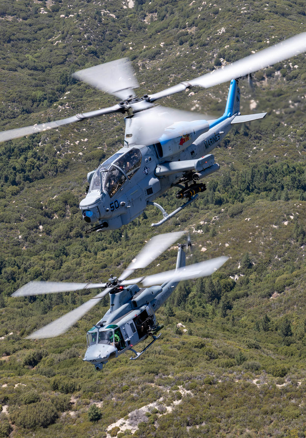 A military helicopter with spinning rotor blades flies over a green mountainous landscape. Description generated by AI