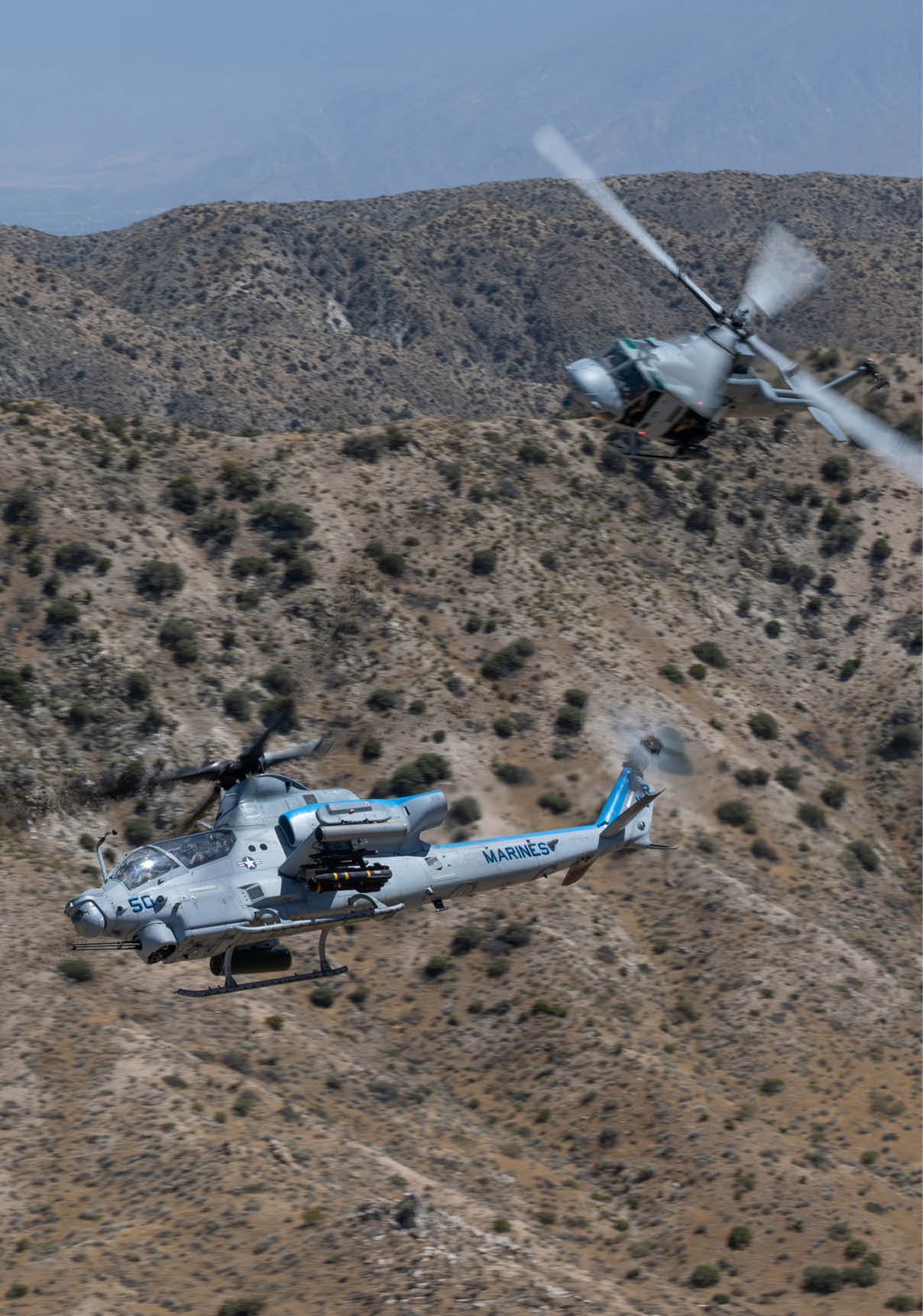 Two military helicopters are flying over a dry, mountainous landscape with sparse vegetation. Description generated by AI