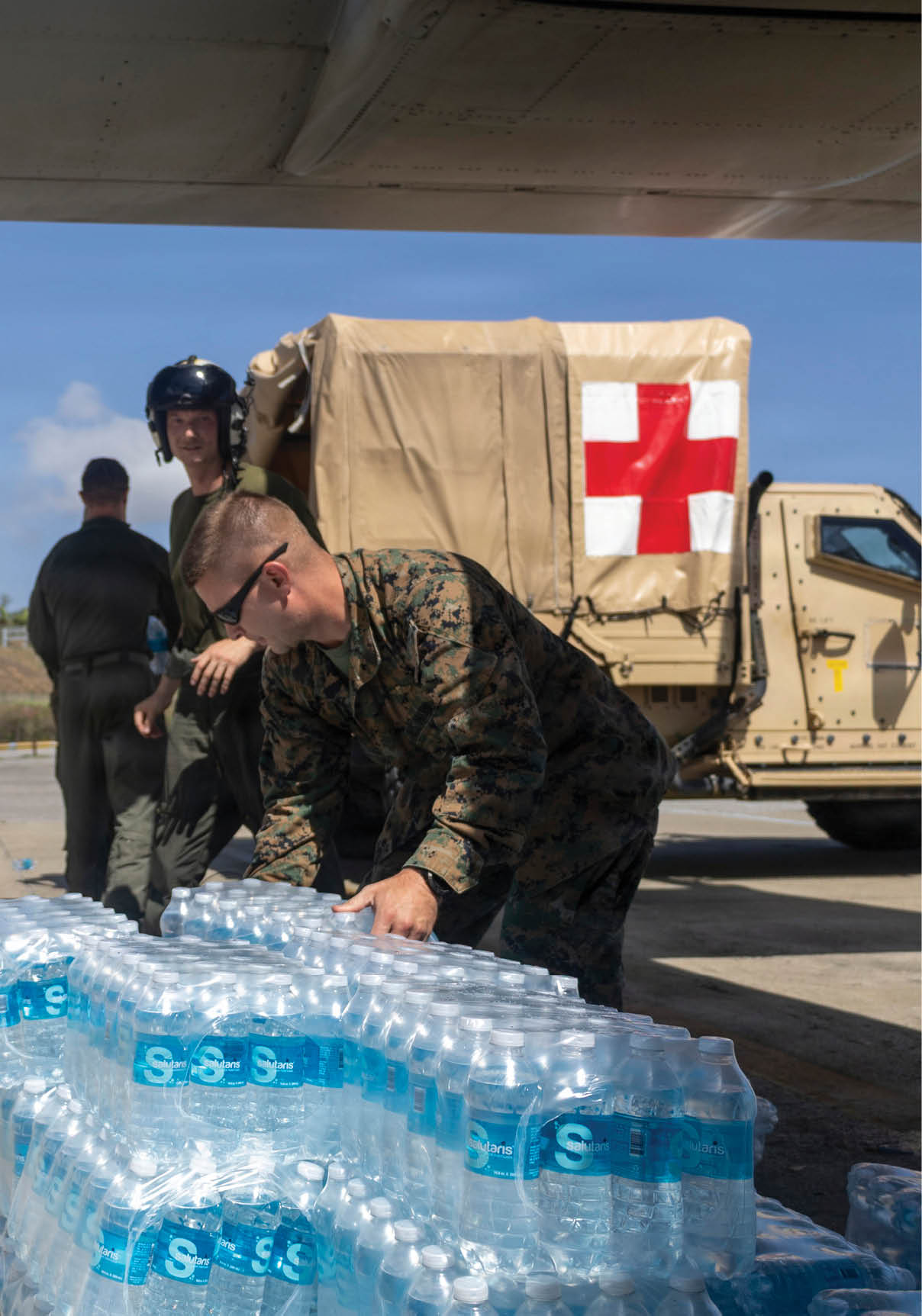 People in military uniforms are unloading bottled water near a vehicle marked with a red cross under the wing of an aircraft. Description generated by AI