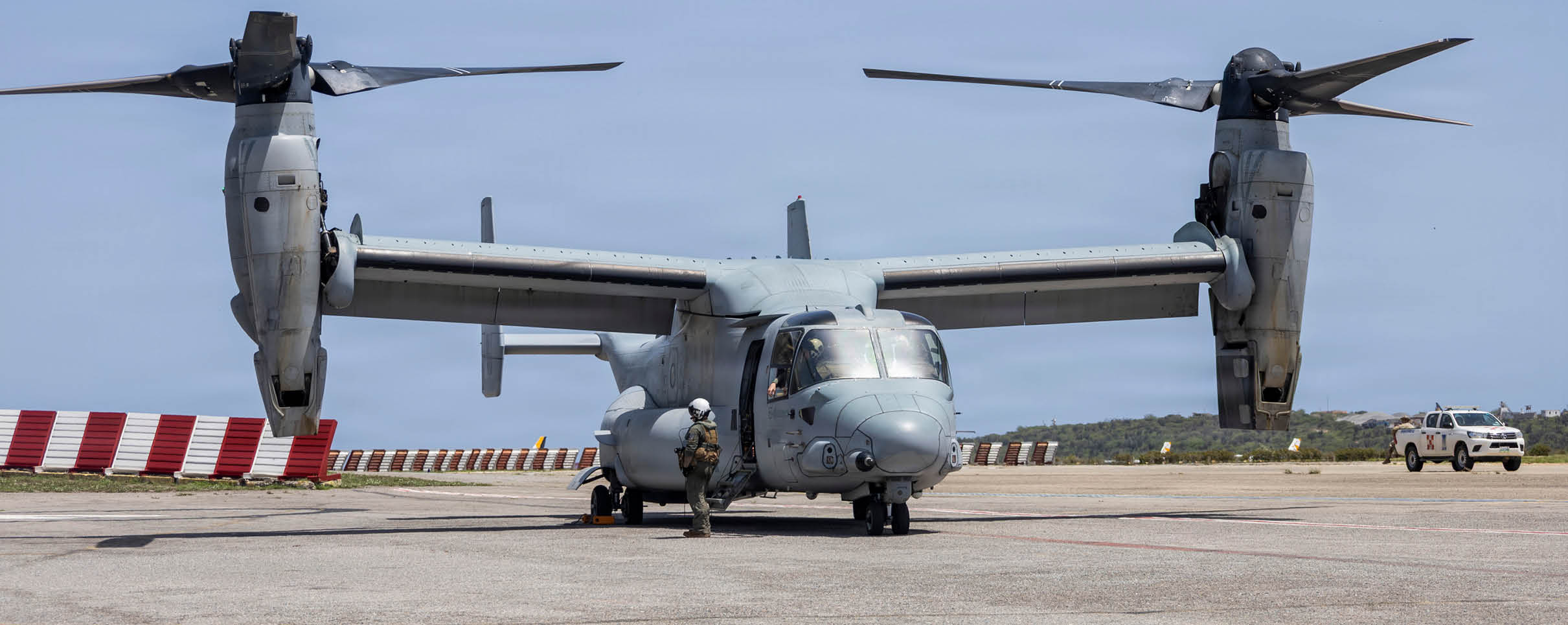 A military tiltrotor aircraft is shown from the front with its rotors positioned horizontally against a clear blue sky. Description generated by AI
