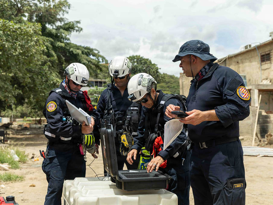Four people wearing helmets and dark uniforms are gathered around an open black case on a white container outdoors near buildings and trees. Description generated by AI