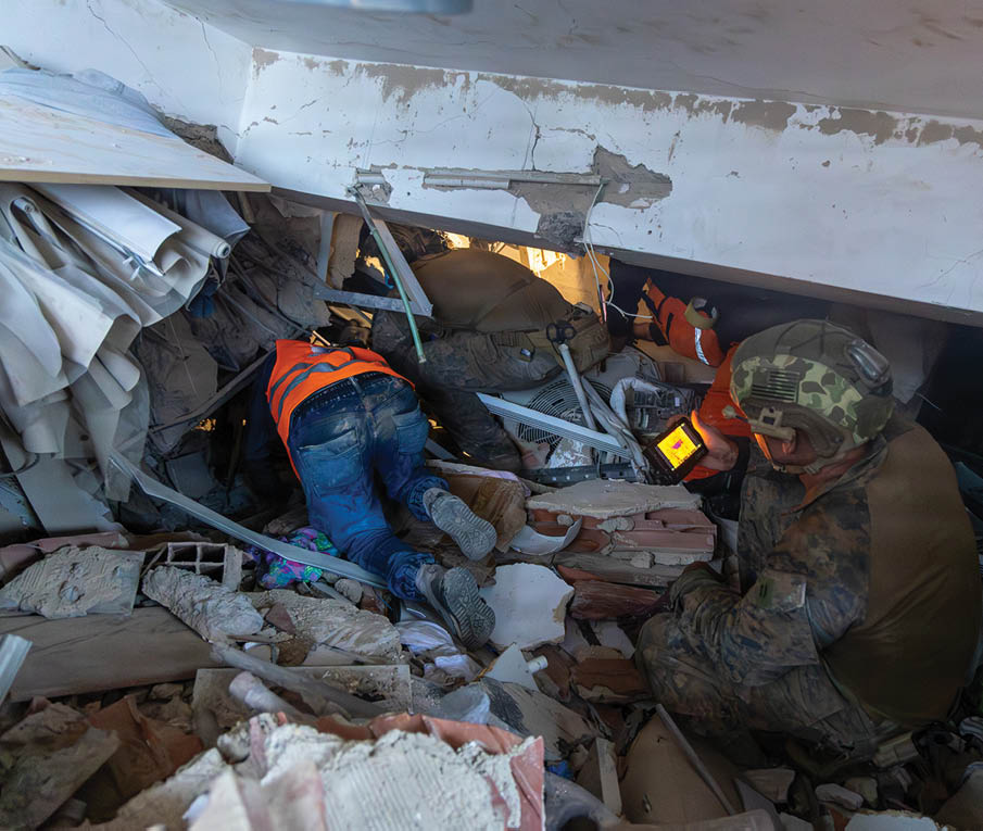 Two people wearing helmets and protective clothing are searching through rubble in a collapsed building, with one person using a flashlight. Description generated by AI