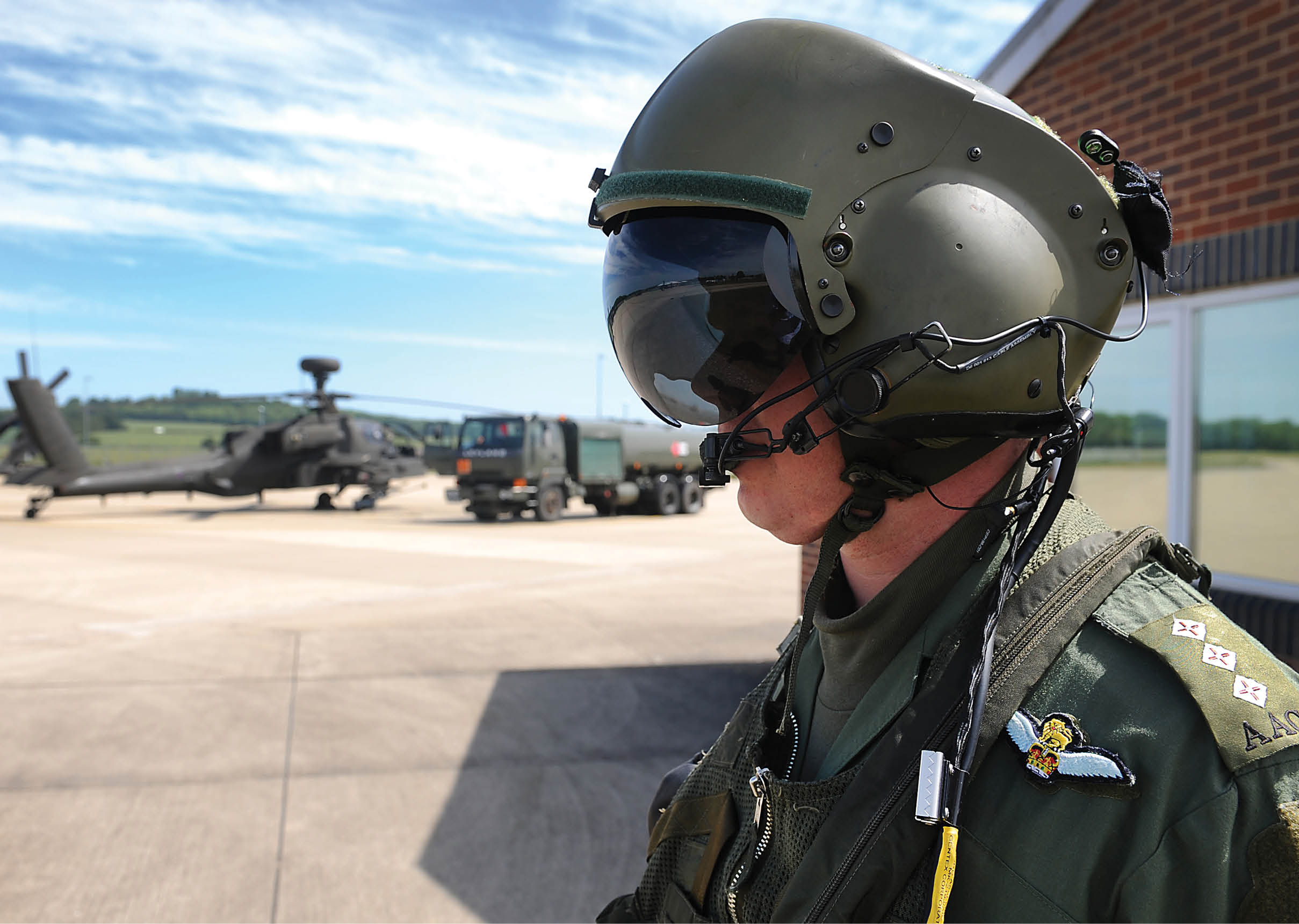 A person wearing a military flight helmet and uniform stands on a tarmac with helicopters and vehicles in the background under a partly cloudy sky. Description generated by AI