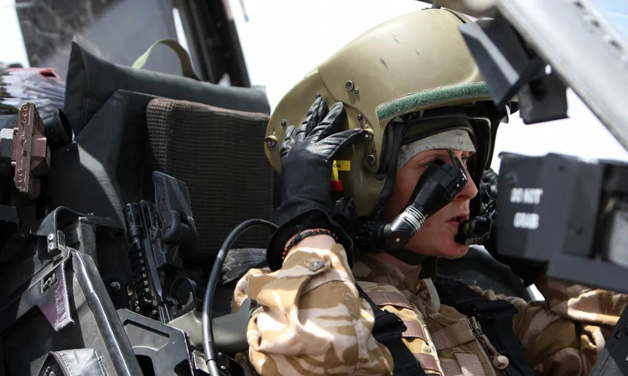 A person wearing a military helmet and camouflage uniform is seated inside a vehicle, adjusting their helmet with one hand near their ear. Description generated by AI