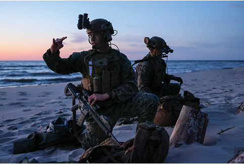 Two men in military uniforms are sitting on the beach, one pointing at the other. They have backpacks and a handbag nearby. AI generated content