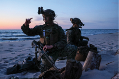 Two men in military uniforms are sitting on the beach, one pointing at the other. They have backpacks and a handbag nearby. AI generated content