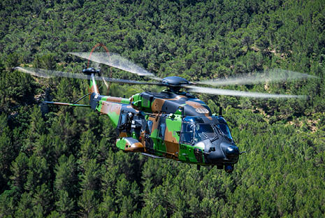 A military helicopter with camouflage paint is flying over a dense forest. Description generated by AI