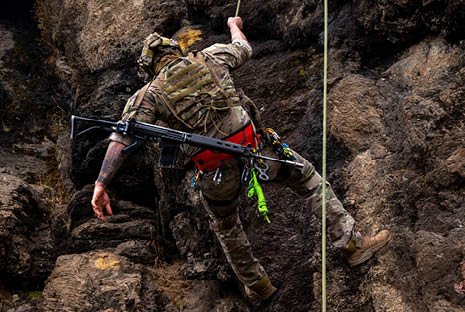 A person wearing camouflage clothing and tactical gear is climbing a rocky surface using a rope while carrying a rifle. Description generated by AI