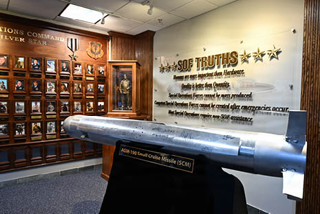 A large missile labeled AGM-190 Small Cruise Missile is displayed on a stand in a museum room with wooden plaques and a glass case containing a military uniform. Description generated by AI