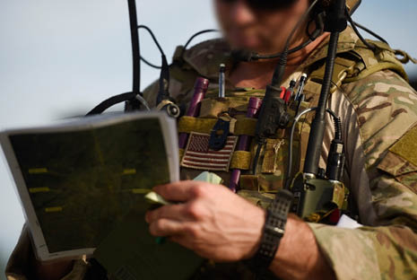A person wearing camouflage military gear and a tactical vest is holding and writing on a map or document outdoors. Description generated by AI