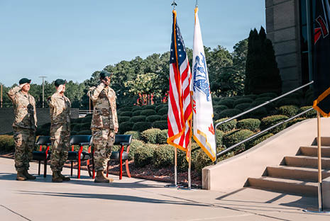 Three adults in military uniforms stand at attention and salute near two flags outside a building with stairs. Description generated by AI