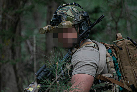A person wearing tactical gear and a camouflage helmet with night vision goggles is holding a device in a forested area. Description generated by AI