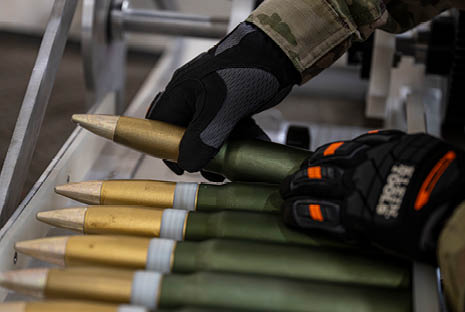 A person wearing gloves and camouflage clothing is handling large artillery shells arranged in a row. Description generated by AI