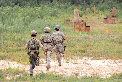 Three people in military-style clothing and helmets are running across a grassy field toward wooden barriers and orange targets. Description generated by AI