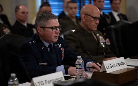 Two adults in military uniforms are seated at a table with microphones and nameplates, appearing to participate in a formal meeting or hearing. Description generated by AI