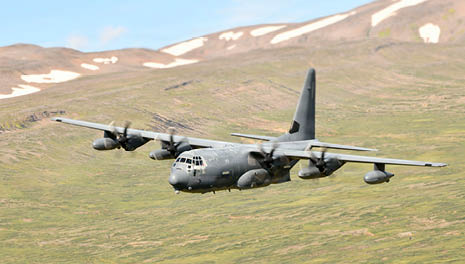 A military cargo plane with four propellers is flying low over a green mountainous landscape with patches of snow. Description generated by AI
