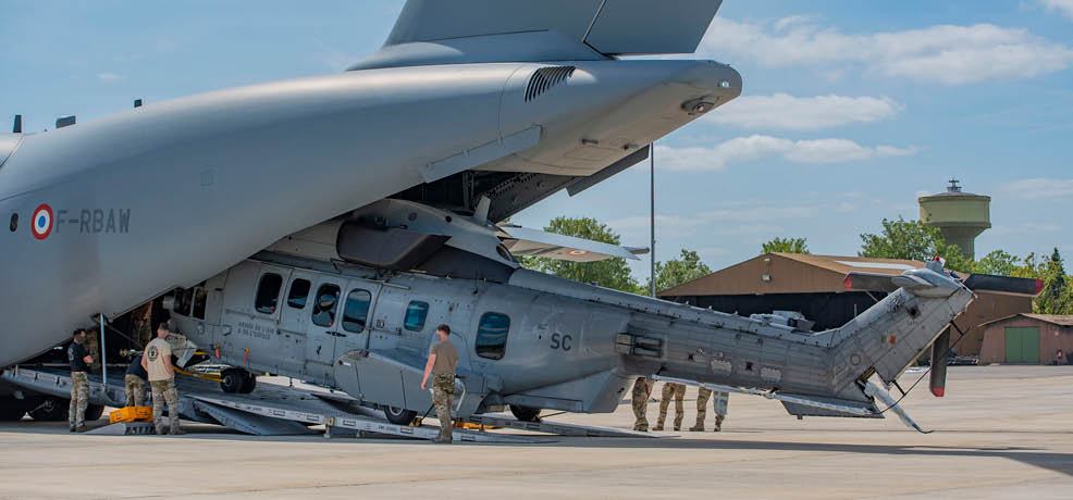 Several people in military uniforms are working around a large helicopter that is partially inside a hangar on a clear day. Description generated by AI
