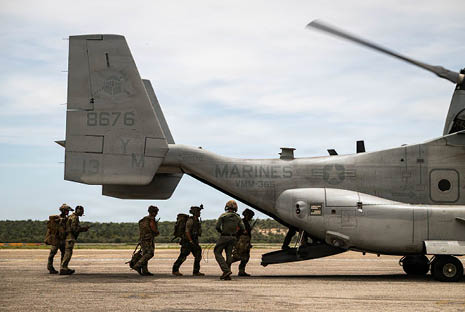 People in military uniforms are walking towards the open rear ramp of a stationary military aircraft on a runway. Description generated by AI