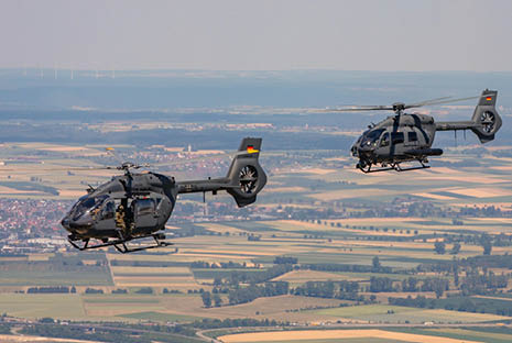 Two military helicopters fly over a rural landscape with fields and scattered buildings visible below. Description generated by AI