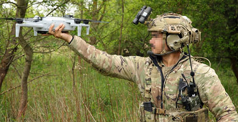 A person wearing camouflage clothing and a tactical helmet is holding a drone in an outdoor wooded area. Description generated by AI